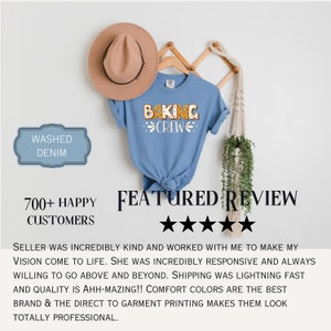 May include: A washed denim blue short-sleeve t-shirt featuring the text BAKING CREW in a gingerbread-style font with white icing details. The shirt is displayed on a wooden hanger alongside a tan fedora hat and a hanging macrame plant holder.