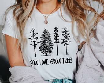 May include: A white short-sleeved t-shirt featuring a black graphic print of four distinct tree silhouettes arranged in a row. Below the trees, the text SOW LOVE, GROW TREES is printed in a bold, black, sans-serif font across the chest area.