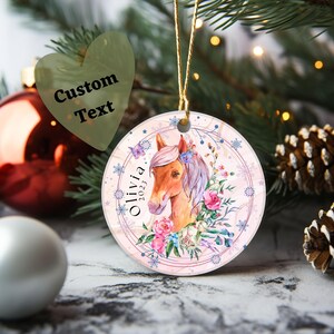 May include: A pink and white Christmas ornament with a watercolor illustration of a brown horse with a pink mane and flowers. The ornament has a gold string and the text "Olivia 2023" is written on the left side.