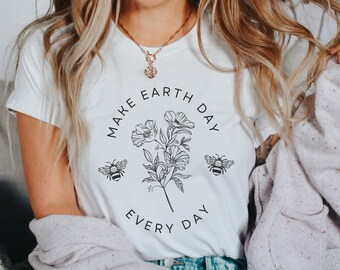 May include: A white short-sleeved t-shirt featuring a black line art graphic of a floral bouquet flanked by two bees. The text "MAKE EARTH DAY EVERY DAY" is printed in a circular arrangement around the central botanical illustration.