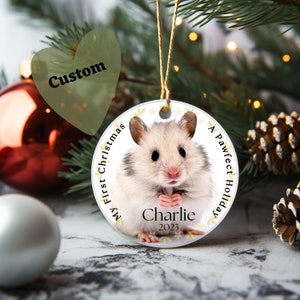 May include: A round Christmas ornament featuring a hamster named Charlie. The ornament has the text "My First Christmas" and "A Pawfect Holiday" around the edge. The year 2023 is also printed on the ornament.