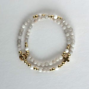 May include: A beaded bracelet featuring smooth, round, off-white and light gray stones interspersed with small, polished gold-tone metal beads. The bracelet includes a circular gold-tone charm with a figure and a small gold-tone cross charm.