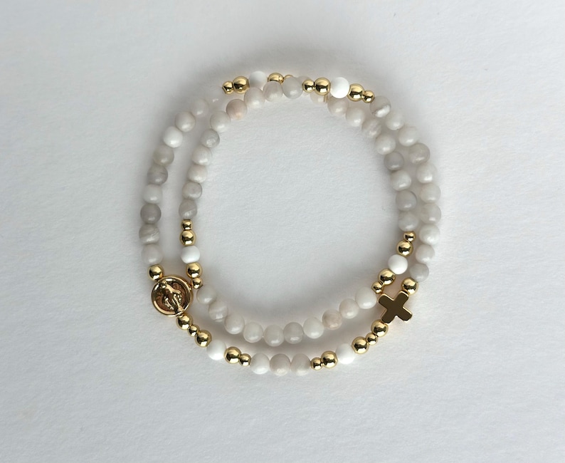 May include: A beaded bracelet featuring smooth, round, off-white and light gray stones interspersed with small, polished gold-tone metal beads. The bracelet includes a circular gold-tone charm with a figure and a small gold-tone cross charm.