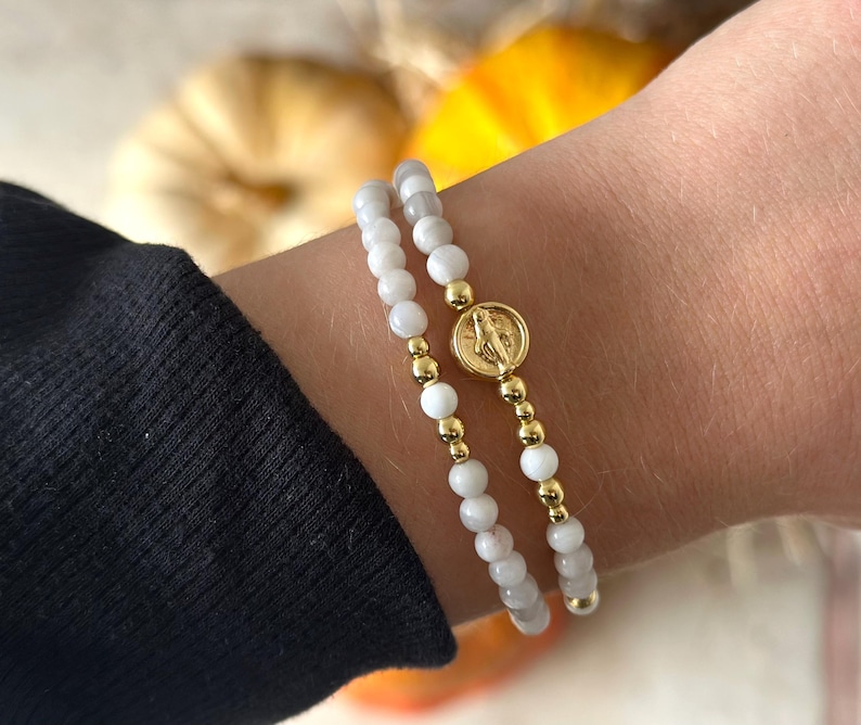 May include: A double-strand bracelet featuring white beads and gold-toned beads, with a central circular gold-toned charm depicting a religious figure. The bracelet is worn on a wrist, with the white beads appearing smooth and round.