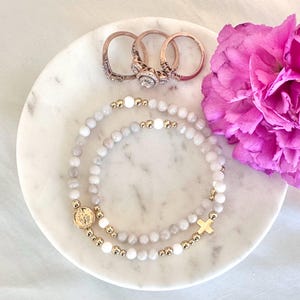 May include: Two beaded stretch bracelets featuring light gray and white stone beads, gold-toned metal spacers, a circular gold-toned charm with an embossed figure, and a gold-toned cross charm, accompanied by three rose gold-toned metal rings.