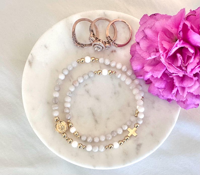 May include: Two beaded stretch bracelets featuring light gray and white stone beads, gold-toned metal spacers, a circular gold-toned charm with an embossed figure, and a gold-toned cross charm, accompanied by three rose gold-toned metal rings.
