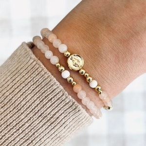 Könnte beinhalten: Zwei Armbänder mit goldenen Perlen und rosafarbenen und weißen Steinen. Ein Armband hat einen goldenen Anhänger mit einem religiösen Bild.