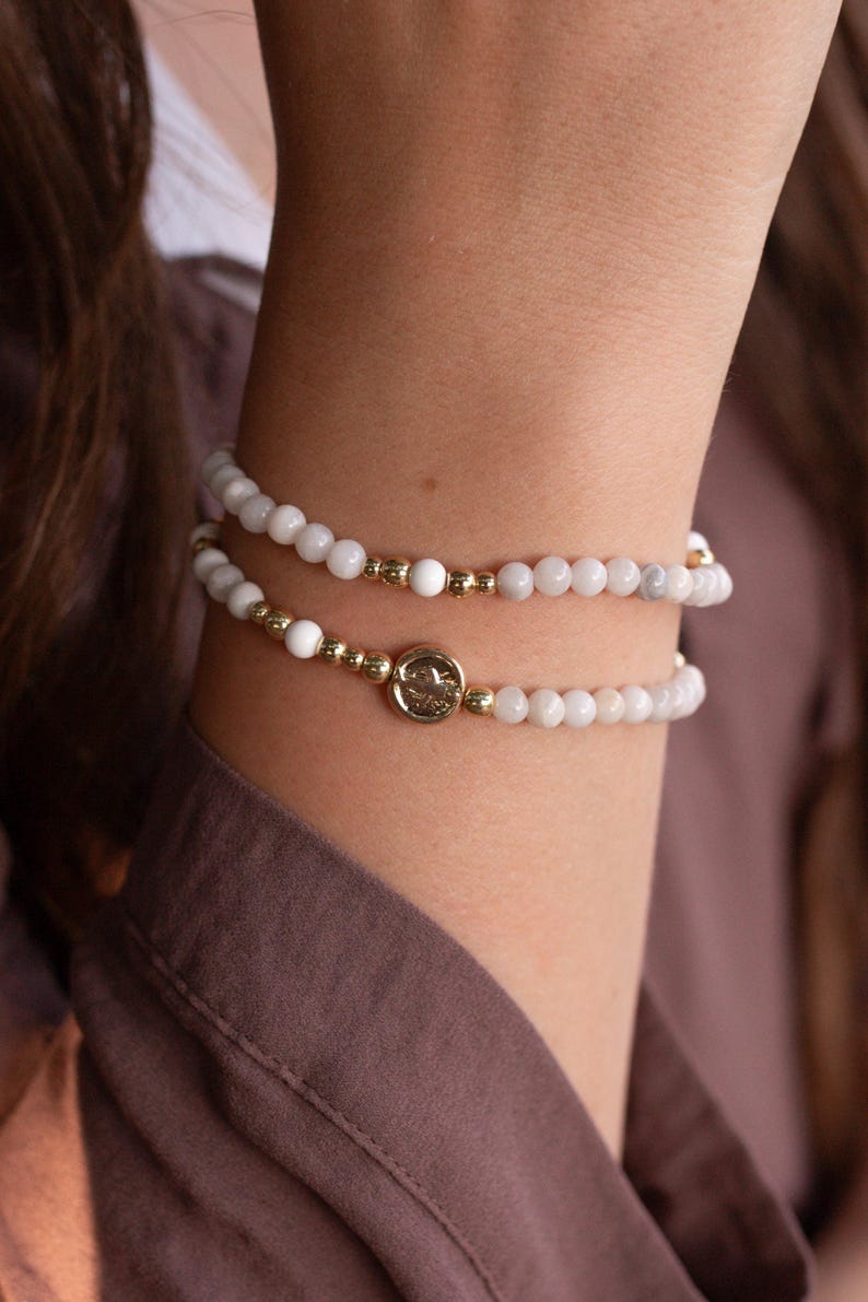 May include: Two beaded bracelets featuring small, smooth white spherical beads interspersed with polished gold-tone metallic beads. One bracelet includes a central circular gold-tone charm with an embossed design, creating a layered look.
