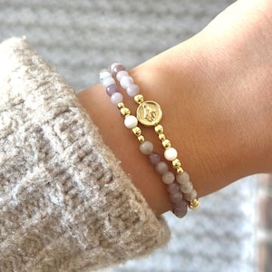Könnte beinhalten: Zwei Armbänder mit goldenen Perlen und einem goldenen Anhänger am Handgelenk einer Person. Die Armbänder bestehen aus hellrosa und weißen Perlen.