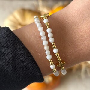 May include: Two beaded bracelets worn together. One bracelet features small white spherical beads with gold-toned accents. The second bracelet consists of white beads interspersed with gold-toned beads and a small gold-toned cross charm.