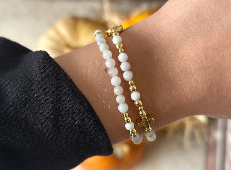 May include: Two beaded bracelets worn together. One bracelet features small white spherical beads with gold-toned accents. The second bracelet consists of white beads interspersed with gold-toned beads and a small gold-toned cross charm.