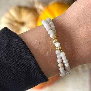 May include: A double-strand beaded bracelet featuring small, smooth, off-white spherical beads. The two strands are connected by a central section consisting of two polished gold-tone metallic beads flanking a single off-white bead.