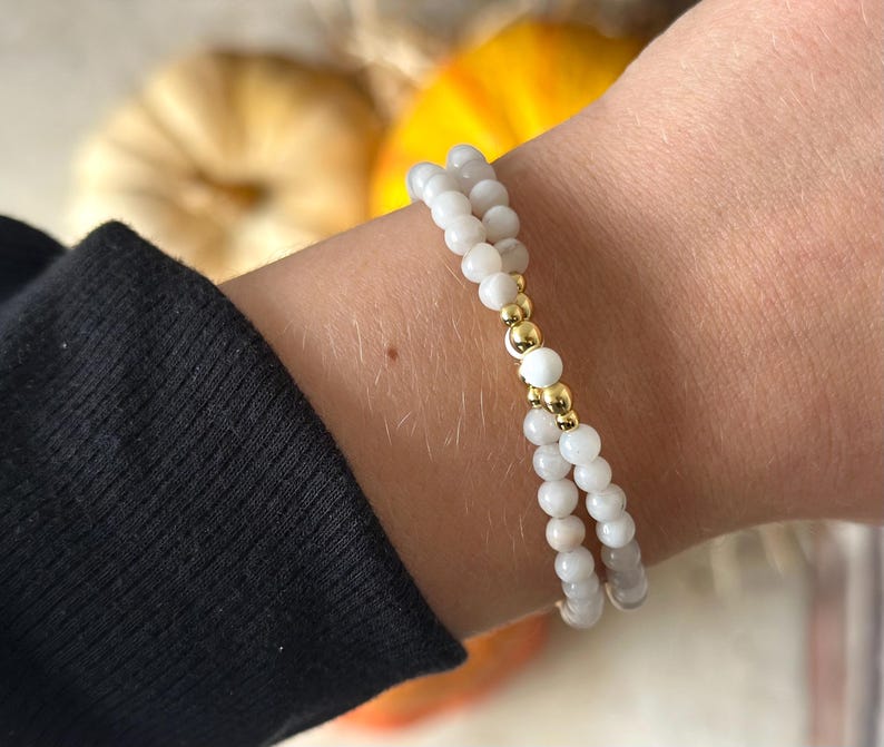 May include: A double-strand beaded bracelet featuring small, smooth, off-white spherical beads. The two strands are connected by a central section consisting of two polished gold-tone metallic beads flanking a single off-white bead.