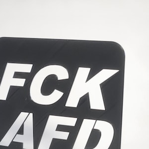 Könnte beinhalten: Schwarzes quadratisches Schild mit weißem Text "FCK AFD" auf einem schwarzen Ständer. Der Ständer trägt den Text "MUHPRINT.DE".