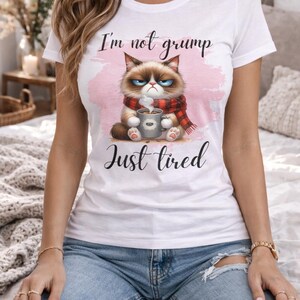 Può includere: T-shirt bianco con un'illustrazione di un gatto scontroso e la scritta "I'm not grump, Just tired." Il gatto indossa una sciarpa rossa e nera e tiene in mano una tazza di caffè. La maglietta è indossata da una persona con jeans blu.