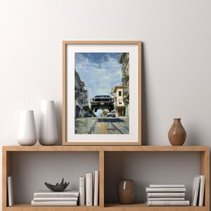 Op de afbeelding: Een ingelijst afdruk van een zwarte auto die over een stadsstraat vliegt. De auto hangt in de lucht, met de voorwielen van de grond. De straat wordt aan beide kanten begrensd door gebouwen, en er zijn bomen en andere auto's op de achtergrond.