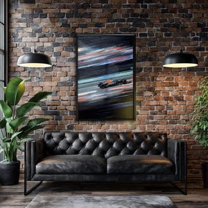 May include: Framed print of a Formula 1 race car in motion, with blurred streaks of color. The artwork is displayed on a brick wall above a black leather sofa. Two black pendant lights hang from the ceiling, and a large potted plant is on the left.