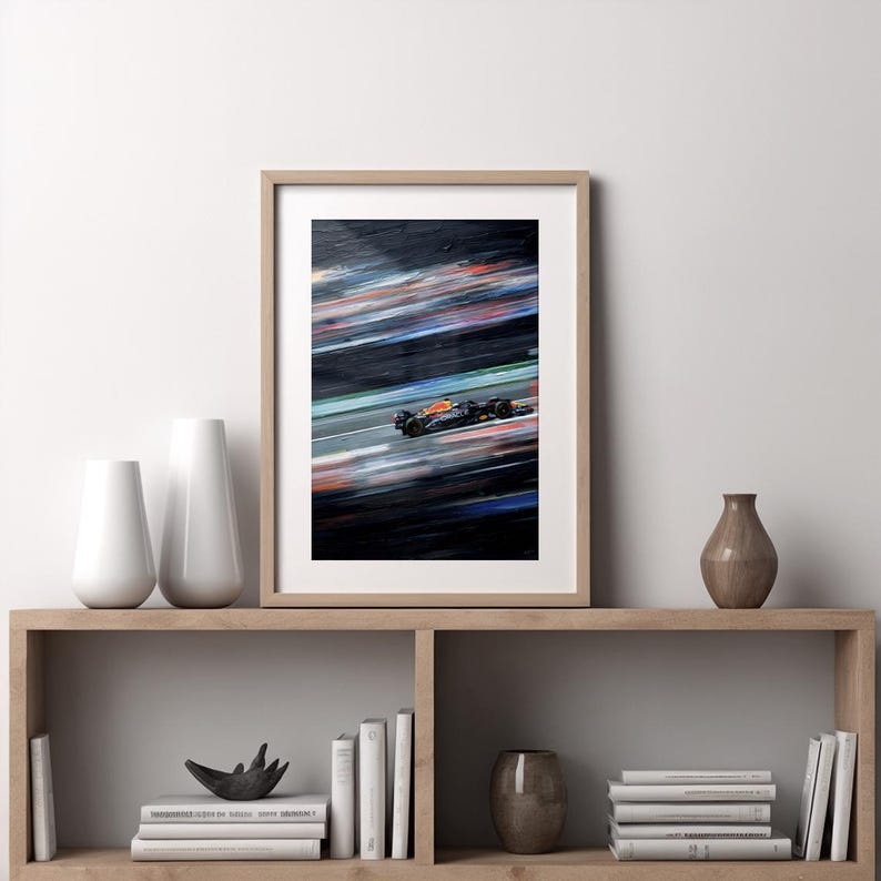 May include: Framed print of a Formula 1 race car in motion. The artwork captures the speed with blurred streaks of color against a dark background. The car is orange and black, set in a light wooden frame, displayed on a wooden shelf.