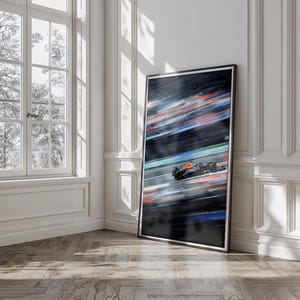 May include: A framed art print depicting a Formula 1 race car in motion. The artwork features a blurred background of colorful streaks, with the car in focus, showcasing its orange, black, and blue design. The print is displayed against a white wall and a wooden floor.