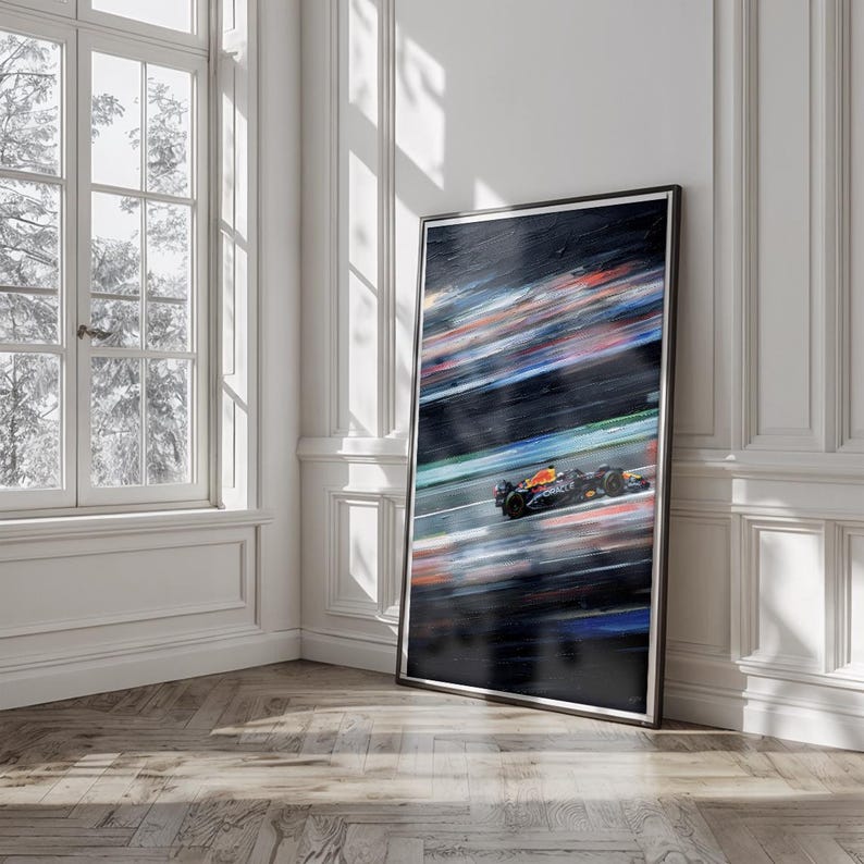 May include: A framed art print depicting a Formula 1 race car in motion. The artwork features a blurred background of colorful streaks, with the car in focus, showcasing its orange, black, and blue design. The print is displayed against a white wall and a wooden floor.