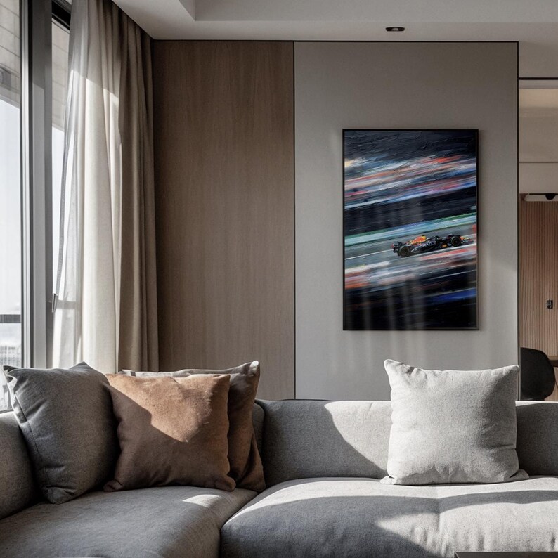 May include: A framed artwork depicting a Formula 1 race car in motion. The painting features streaks of color, including red, blue, and orange, suggesting speed and motion. The artwork is displayed above a gray sectional sofa with several pillows.