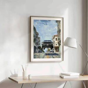Op de afbeelding: Een ingelijst schilderij van een groene auto die over een straat in San Francisco vliegt. De auto is een klassieke Ford Mustang. Het schilderij is in een zilveren lijst en hangt aan een witte muur.