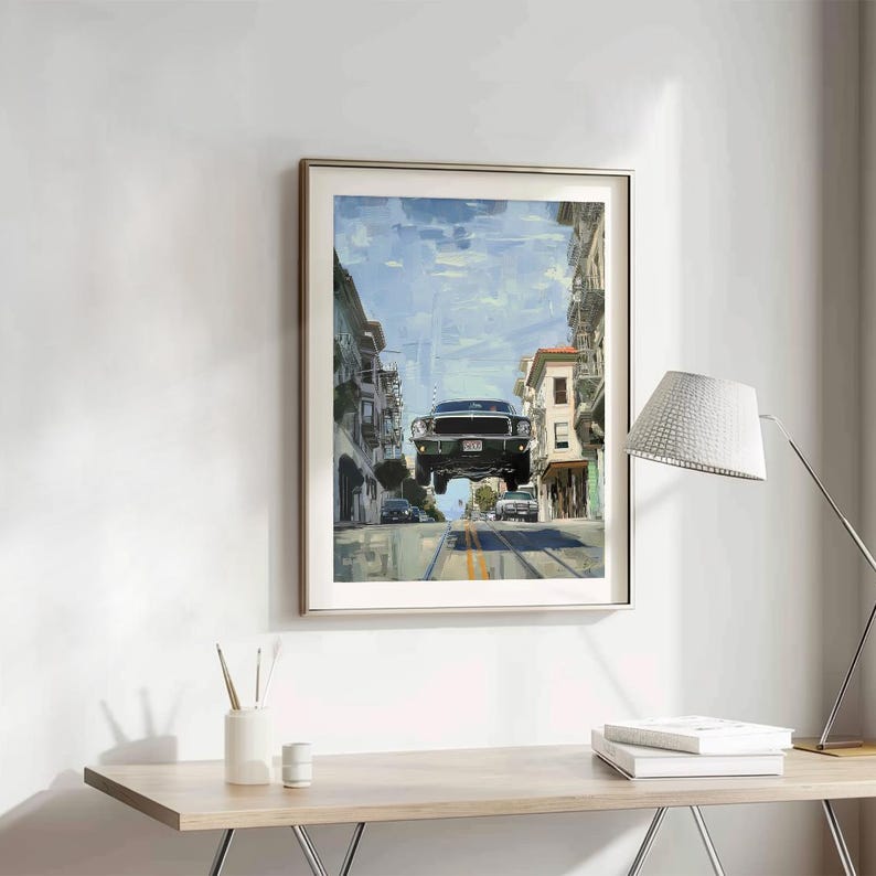 Op de afbeelding: Een ingelijst schilderij van een groene auto die over een straat in San Francisco vliegt. De auto is een klassieke Ford Mustang. Het schilderij is in een zilveren lijst en hangt aan een witte muur.