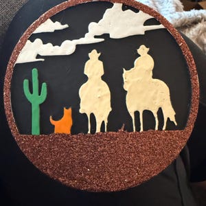 May include: A round, decorative ornament featuring a Western scene. Two riders on horseback are silhouetted against a black background, with a cactus and an orange cat. The border is a glittery brown, and the clouds are white.