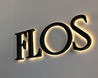 May include: A three-dimensional wall sign displaying the word FLOS in bold, black, serif capital letters. Each letter is mounted slightly away from the wall, creating a soft, warm, glowing halo effect behind the characters from integrated lighting.
