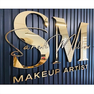 Puede incluir: Letrero dorado y espejado con el texto "SM Sarah Mellow MAKEUP ARTIST" sobre una pared azul oscuro con líneas verticales. Las letras doradas reflectantes crean un efecto visual llamativo. Para una maquilladora.