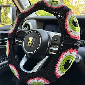 May include: A black steering wheel cover with a crocheted design featuring pink, green, and white eye patterns.