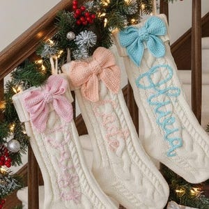 Peut inclure: Trois bas de Noël en tricot de couleur crème avec des nœuds roses, pêche et bleus sont suspendus à un escalier en bois. Chaque bas porte un nom brodé dans une couleur assortie. L'escalier est décoré de guirlandes, de lumières et de cadeaux.