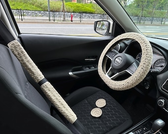 May include: Beige crocheted steering wheel cover and seatbelt cover with two beige crocheted coasters in a car interior.