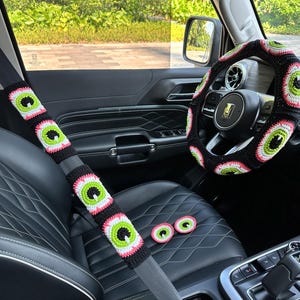 May include: A black car interior with a crocheted steering wheel cover and seatbelt covers featuring a repeating eye pattern. The covers are black, pink, and green.