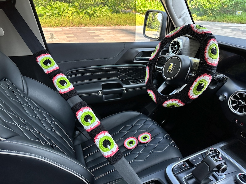 May include: A black car interior with a crocheted steering wheel cover and seatbelt covers featuring a repeating eye pattern. The covers are black, pink, and green.