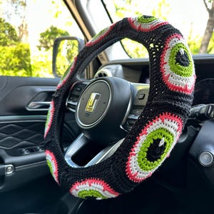 May include: A black, white, green, and pink crocheted steering wheel cover with a repeating eye design.