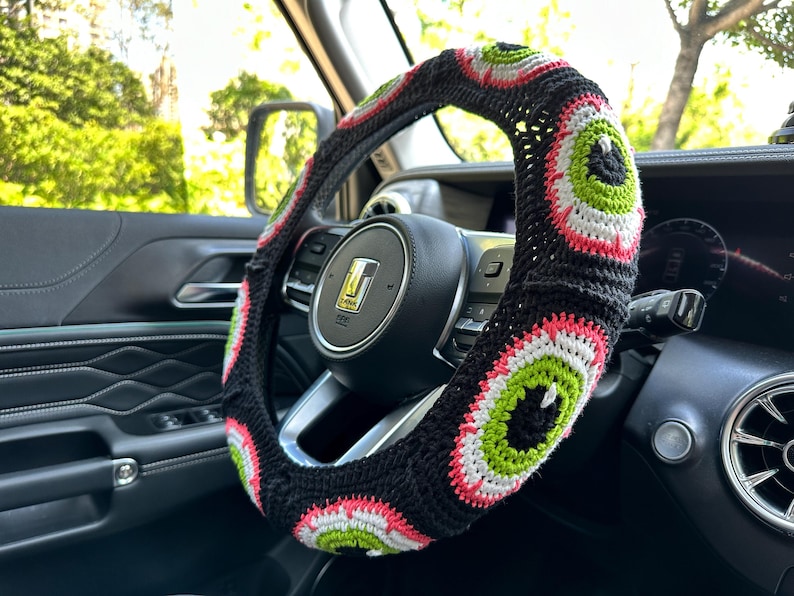 May include: A black, white, green, and pink crocheted steering wheel cover with a repeating eye design.