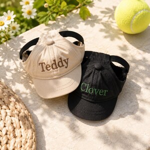 May include: Two pet baseball caps with ear holes, one beige with brown embroidery reading Teddy and one black with green embroidery reading Clover. Each cap features a structured crown, a curved brim, and a black adjustable strap for secure fit.
