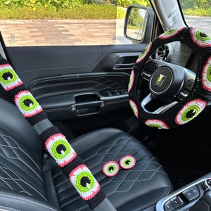 May include: A black car interior with a crocheted steering wheel cover and seatbelt covers featuring a green eye design. There are also three small crocheted eye-shaped coasters on the center console.