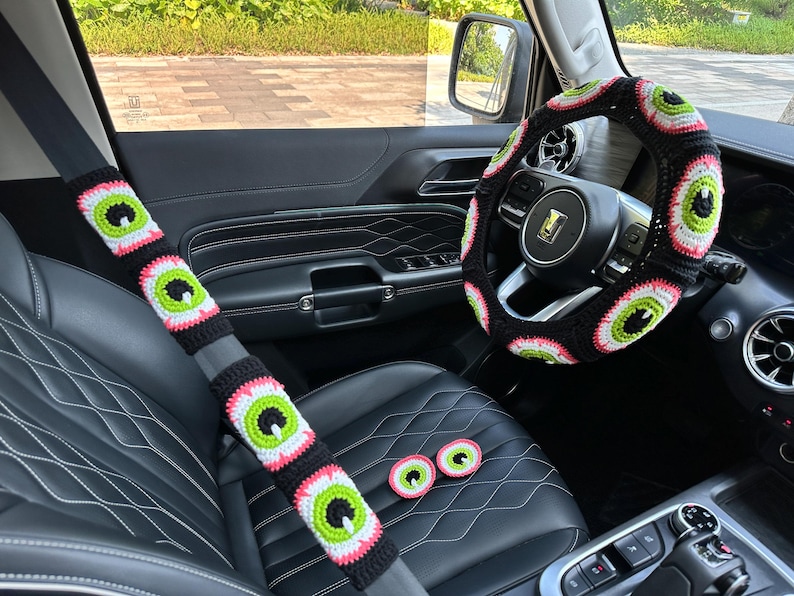 May include: A black car interior with a crocheted steering wheel cover and seatbelt covers featuring a green eye design. There are also three small crocheted eye-shaped coasters on the center console.