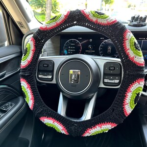 May include: A black steering wheel cover with a colorful crocheted design. The cover features a repeating pattern of green, white, and pink circles.
