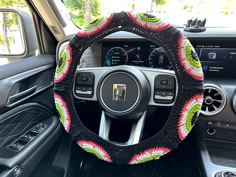 May include: A black steering wheel cover with a colorful crocheted design. The cover features a repeating pattern of green, white, and pink circles.
