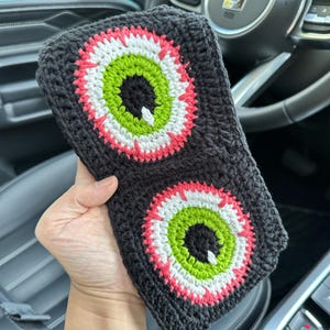 May include: A black crocheted headband with two large, colorful eyes. The eyes are green, white, and pink. The headband is in a car.