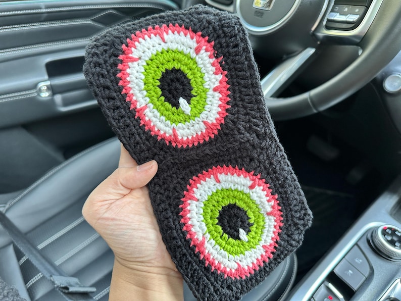 May include: A black crocheted headband with two large, colorful eyes. The eyes are green, white, and pink. The headband is in a car.