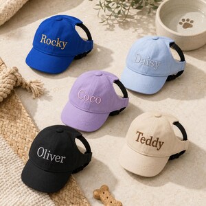 May include: Five baseball caps for pets in blue, light blue, purple, black, and beige. Each cap features an adjustable black strap and is embroidered with a name: Rocky, Daisy, Coco, Oliver, or Teddy. The caps have ear holes on the sides.