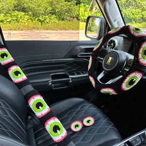 May include: A black car interior with a crocheted steering wheel cover and seatbelt covers. The covers feature a repeating pattern of green, pink, and white eyes.