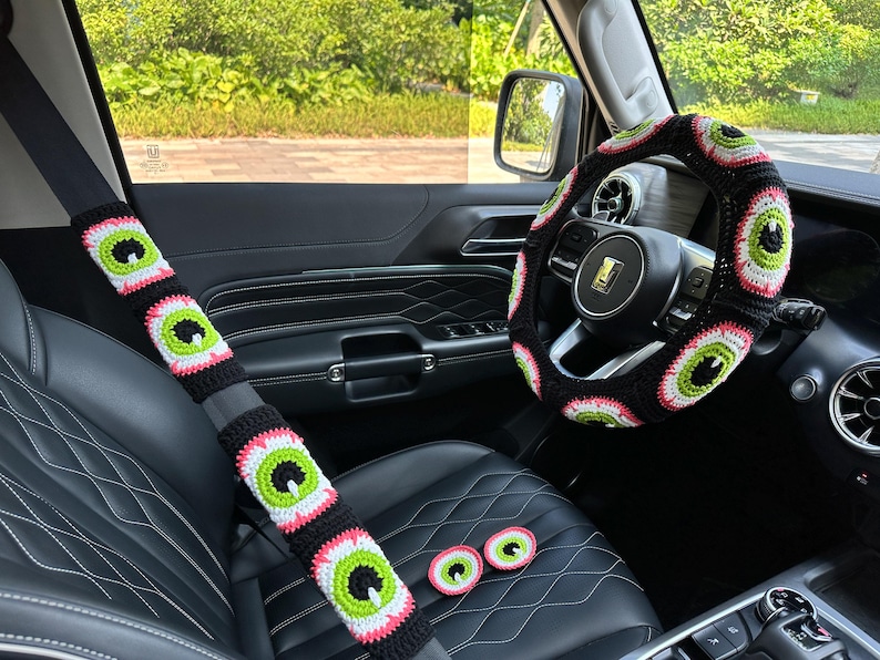 May include: A black car interior with a crocheted steering wheel cover and seatbelt covers. The covers feature a repeating pattern of green, pink, and white eyes.