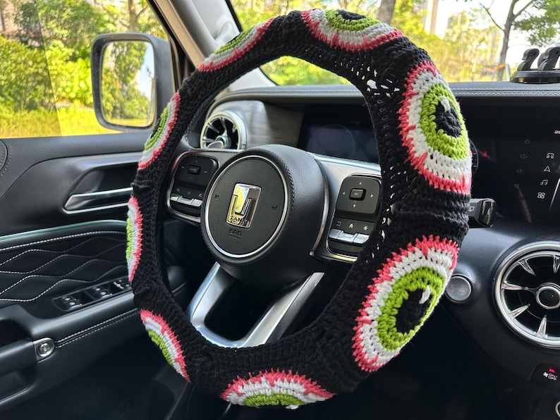 May include: A black steering wheel cover with a crocheted design featuring pink, green, and white eye patterns.