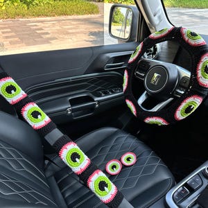 May include: A black car interior with a crocheted steering wheel cover, seatbelt covers, and headrest covers. The covers are all black with green and pink eye designs.