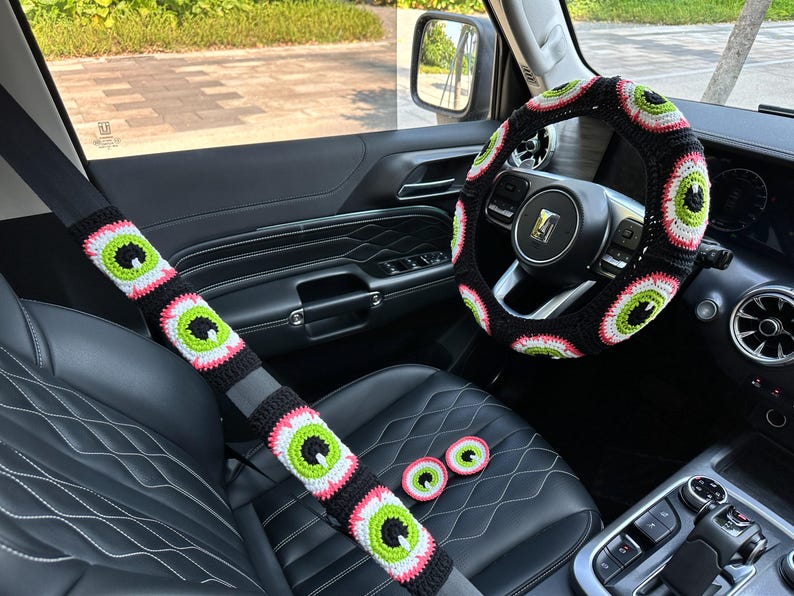 May include: A black car interior with a crocheted steering wheel cover, seatbelt covers, and headrest covers. The covers are all black with green and pink eye designs.
